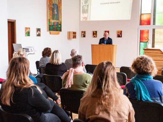 Ein Mann steht an einem Rednerpult und hält eine Präsentation vor einem Publikum. Im Hintergrund ist eine Projektionsleinwand zu sehen, und an den Wänden hängen religiöse Bilder und Kunstwerke.