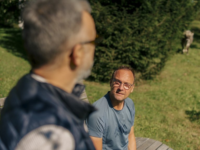 Ein Mann sitzt auf einer Bank auf einer Wiese, ein anderer Mann steht daneben.